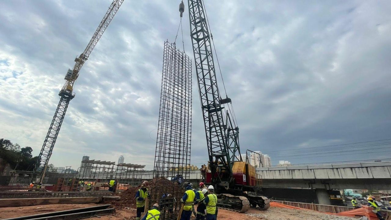 Dois guindastes sobre esteiras posicionam a armação de um pilar enquanto a equipe trabalha na base, em obra urbana de infraestrutura.