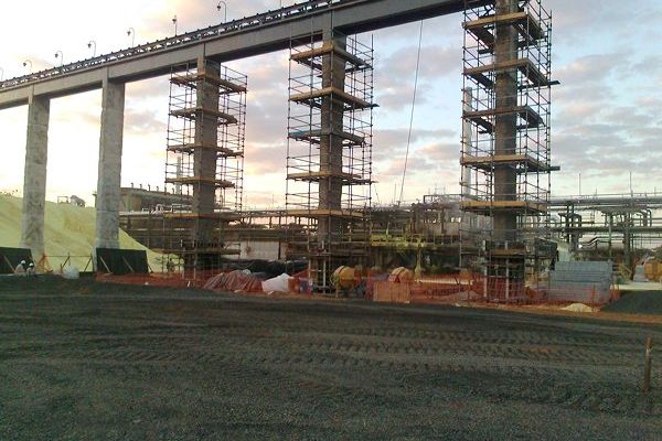 Estrutura elevada de concreto em execução, com pilares, escoramento e andaimes, ao lado de via existente.