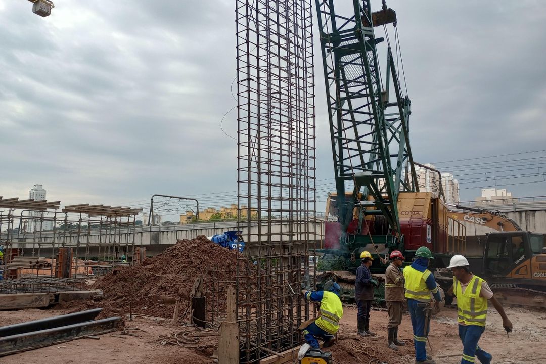Equipe com capacete e colete de alta visibilidade executa a armação de fundação ao lado de guindaste e escavadeira.