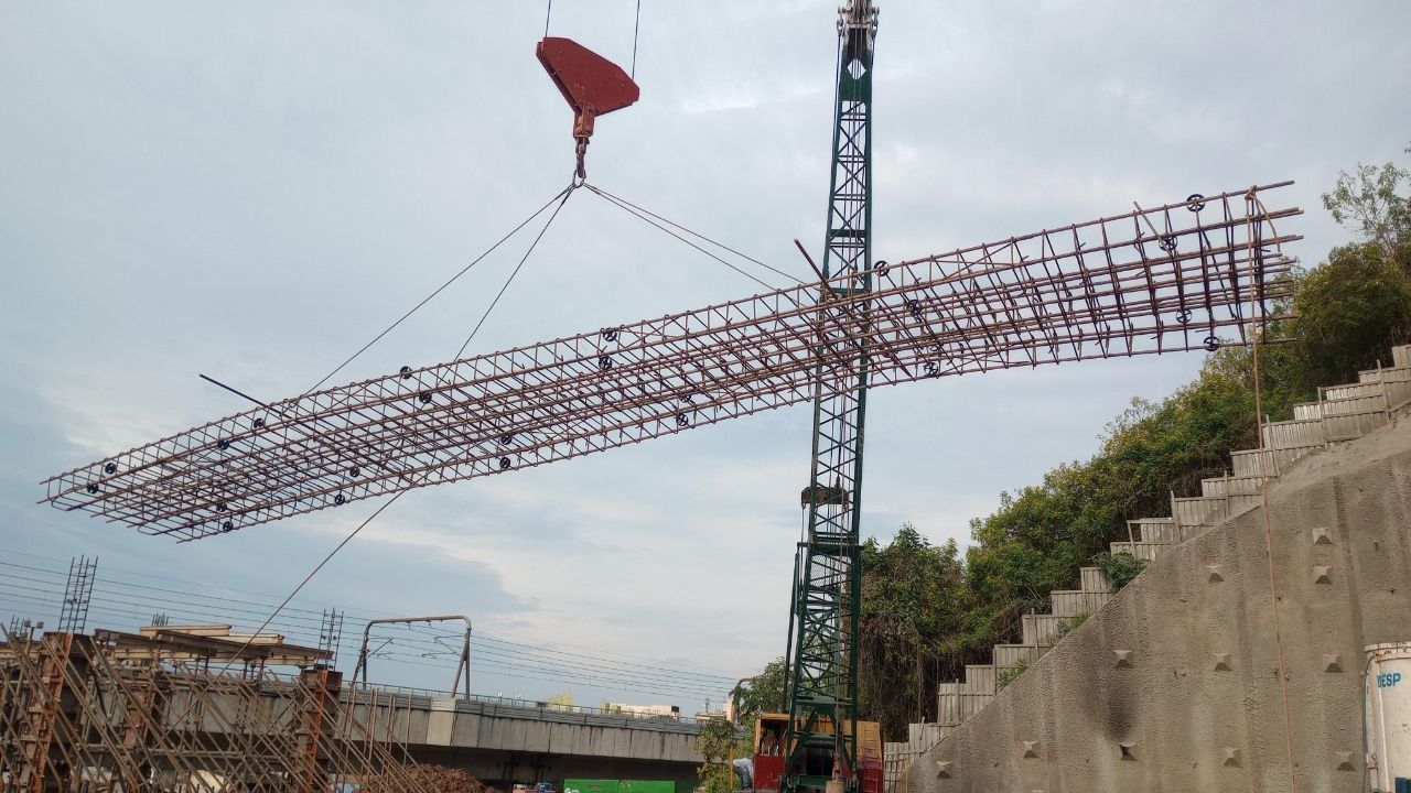 Guindaste iça uma armadura longitudinal de concreto armado ao lado de uma contenção em talude.