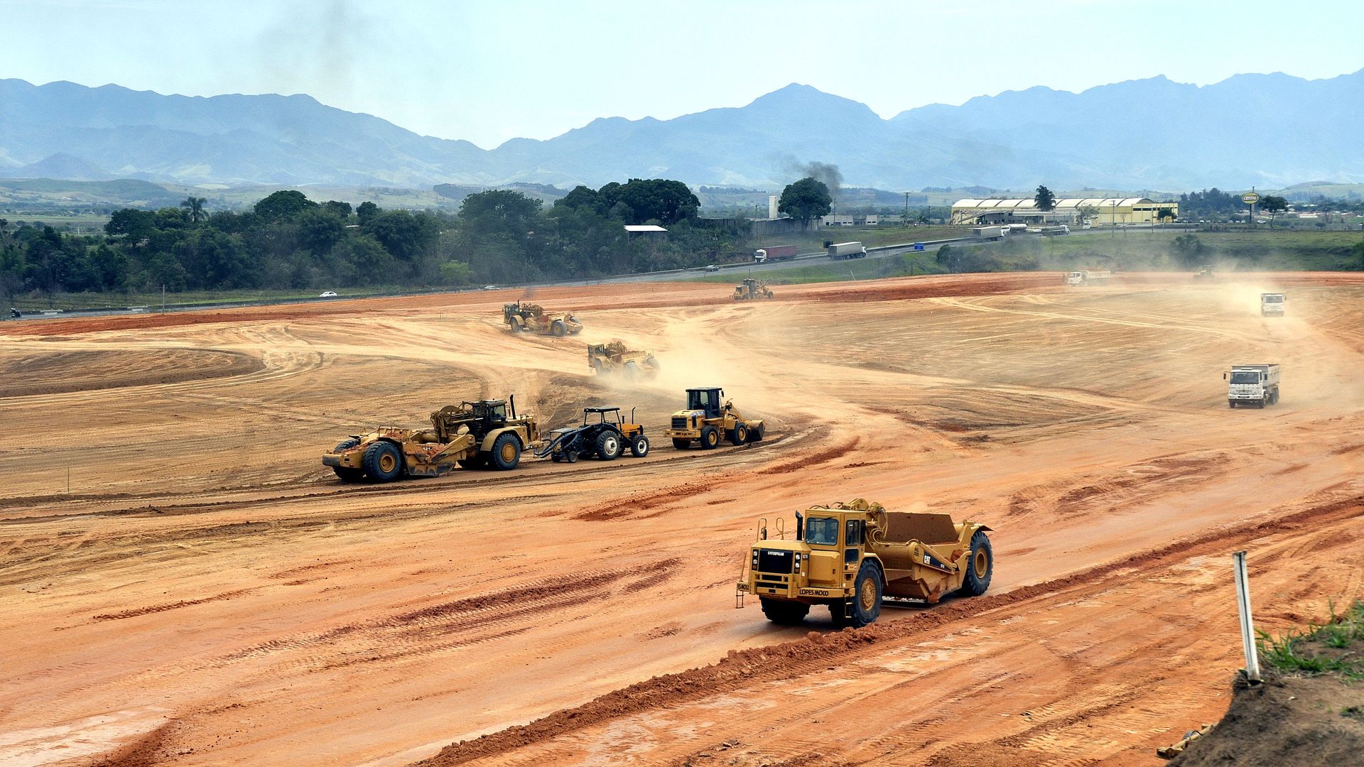 Frente ampla de terraplenagem com scrapers, pá carregadeira e caminhões movimentando solo, com serra ao fundo.